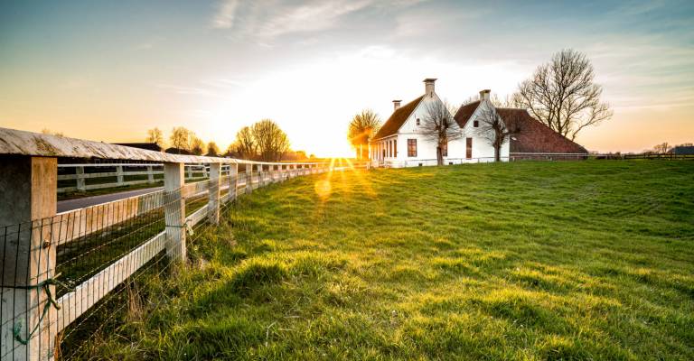 Landweggetje in het Nederlandse landschap met wit hek dat leidt naar een oude boerderij in Nederland tijdens een sfeervolle zonsondergang